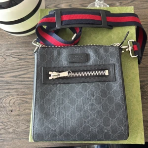 Axelväska från Gucci - Snygg axelväska från Gucci i mörkgrått med diskret mönster och en framficka med dragkedja. Väskan har en justerbar axelrem i rött och blått, perfekt för en stilren look. Priset går att diskuteras hör av er !