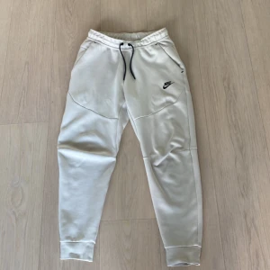 Nike tech fleece byxor, Beige - Säljer ett par Nike tech fleece byxor med elastisk midja och snörning. Byxorna har sidofickor och en liten Nike-logga på ena benet. Perfekta för en avslappnad stil.