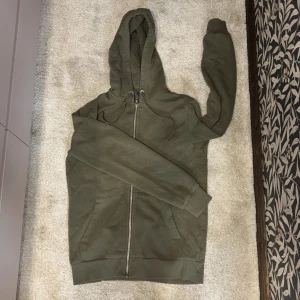 Olivgrön hoodie med dragkedja - Säljer en olivgrön hoodie med dragkedja och huva. Tröjan har långa ärmar och ribbade muddar vid ärmslut och nederkant. 