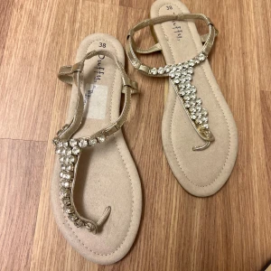 Guldiga sandaler med strassdetaljer - Snygga guldiga sandaler från Blowfish med glittrande strassdetaljer på remmarna. Perfekta för sommarens alla tillfällen. Sandalerna har en bekväm passform och en stilren design.
