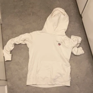 Vit hoodie från GANT - Säljer en klassisk vit hoodie från GANT med en känguruficka framtill och broderad logotyp på bröstet. Perfekt för en avslappnad stil. Hör av dig vid intresse!