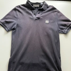 Grå pikétröja från Stone Island - Säljer en stilren grå pikétröja i storlek L slim fit från Stone Island. Tröjan har en klassisk krage med knappar och en diskret logotyp på bröstet. 