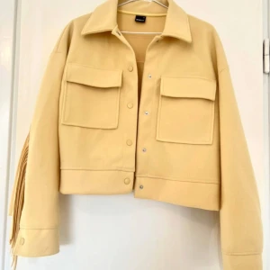 Beige jacka med fransar - Snygg beige jacka med fransar på baksidan och ärmarna. Jackan har en klassisk krage och knappar framtill. Två stora fickor på bröstet ger en cool look. Perfekt för att sticka ut i mängden!