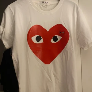 CDG T-shirt - Säljer en vit t-shirt från Comme des Garçons med ett stort rött hjärta med ögon på framsidan. T-shirten är kortärmad och gjord i mjuk bomull. Perfekt för en stilren och lekfull look! 