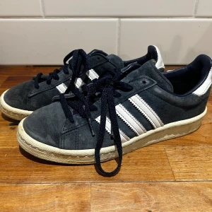 Marinblå adidas campus 80s låga - Snygga marinblå sneakers från Adidas med vita detaljer och snörning. Skorna har en klassisk design med tre ränder på sidan och en bekväm sula. Perfekta för en stilren look.