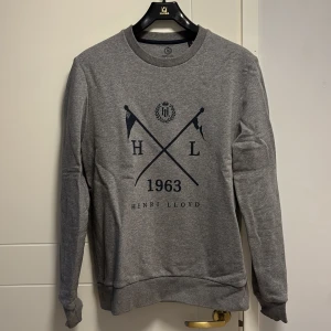 Grå sweatshirt från Henri Lloyd - Säljer en stilren grå sweatshirt från Henri Lloyd med tryck framtill. Tröjan har långa ärmar och rund halsringning. Perfekt för en avslappnad stil.