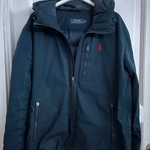 Polo Ralph Lauren jacka - Marinblå ralph lauren jacka i okej skick. Jackan är tunnare och passar bra till vår/sommar. Dragkedjorna på sidorna funkar men kan behövas fixas med själva dragdelen för enklare användning