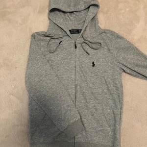 Grå zip hoodie från Polo Ralph Lauren - Stilren och tidlös zip-hoodie i mjuk bomullsblandning, perfekt för en avslappnad men ändå sofistikerad look. Försedd med dragkedja, justerbar huva och broderad signaturlogga på bröstet. Ett mångsidigt plagg som fungerar året runt. 