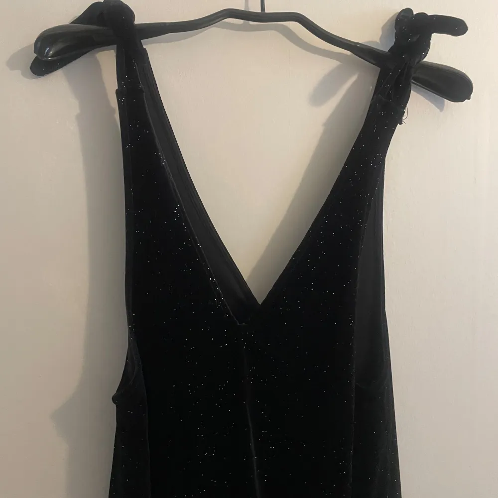 Elegant svart jumsuit med glittrande detaljer. Klänningen har en djup v-ringning och är ärmlös, vilket ger en stilren och festlig look. Perfekt för speciella tillfällen där du vill glänsa.. Mekot.
