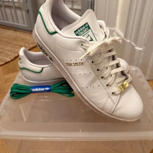 Adidas Stan Smith i vitt och grönt - Klassiska Adidas Stan Smith sneakers i vitt med gröna detaljer. Skorna har en stilren design med snörning och guldtext på sidan. Perfekta för en avslappnad stil. Storlek 45