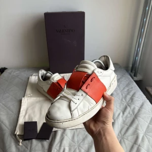 Valentino Garavani sneakers med röd detalj - Snygga Valentino Garavani sneakers i vitt med en iögonfallande röd detalj över snörningen. Skorna har en klassisk design med en modern twist och är perfekta för att lyfta vilken outfit som helst.