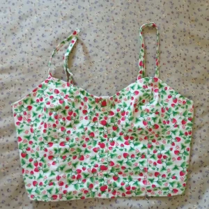 Somrig linnetopp från H&M - Säljer en söt somrig linnetopp från H&M med smala axelband och ett färgglatt mönster av jordgubbar/smultron med rosa blommor och även gröna löv på vit bakgrund. Perfekt för sommardagar! 🌸🍓 Skriv gärna om du vill ha mer information om toppen.😊