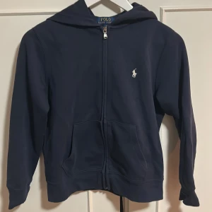 Mörkblå hoodie från Polo Ralph Lauren - Säljer en stilren mörkblå hoodie från Polo Ralph Lauren med dragkedja och klassisk logga på bröstet. Storlek 10-12 år. Tröjan har långa ärmar och en praktisk huva. Perfekt för en avslappnad look.