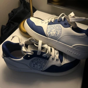 Blå och vita sneakers från Versace - Snygga sneakers från Versace i blått och vitt med det ikoniska Medusa-huvudet på sidan. Skorna har snörning och en bekväm sula med Versace-loggan på hälen. Perfekta för en stilren look. Skorna är köpta i Barcelona nypris 4000kr
