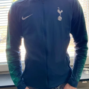 Blå träningsjacka från Nike - Säljer en blå träningsjacka från Nike med Tottenham Hotspur-logga på bröstet. Jackan har långa ärmar med gröna detaljer och en dragkedja framtill. Perfekt för fotbollsträning eller som en sportig vardagsjacka.