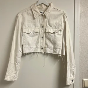Vit croppad jeansjacka från Zara - Säljer en vit croppad jeansjacka från Zara med långa ärmar och knappar framtill. Jackan har två bröstfickor med knappdetaljer och en rå kant nedtill för en trendig look.