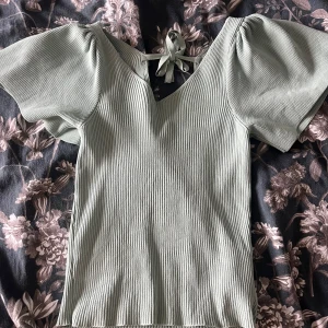 Grön ribbad topp med knytband - Snygg grön ribbad topp med korta puffärmar och v-ringning. Toppen har ett knytband i ryggen som ger en fin detalj. Perfekt för en stilren look.