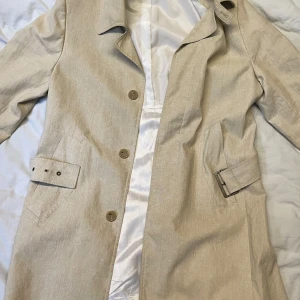 Beige trenchcoat från Tiger of Sweden - Snygg beige trenchcoat från Tiger of Sweden med klassisk design. Jackan har knappar framtill och ett bälte i midjan för en stilren look. Perfekt för vår och höst! Mycket bra skick och sparsamt använd dock en liten skavank. En knapp har ramlat av men medkommer i köpet så man kan laga själv. Osäker på vilken storlek den är men passar som S /M