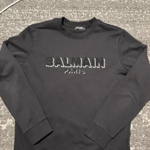 Balmain - Säljer en stilren svart långärmad tröja från Balmain med broderad logotyp på bröstet. Perfekt för en trendig och avslappnad look. Tröjan har en rund halsringning och är tillverkad i ett mjukt material.