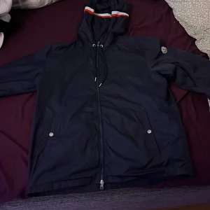 Mörkblå jacka från Moncler - Säljer en snygg mörkblå jacka från Moncler med huva och dragkedja. Jackan har  två fickor med knappar framtill. Detaljer i form av en logotyp på ärmen och en randig kant på huvan.