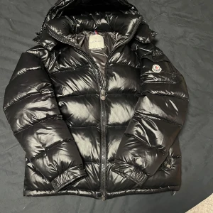 Moncler Maya  - Säljer en snygg svart pufferjacka från Moncler med hög krage och dragkedja. Jackan har en quiltad design och en broderad logotyp på ärmen. Perfekt för kyliga dagar med sin varma och stilrena look.