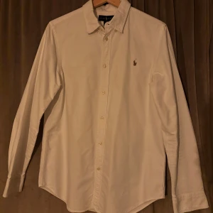 Vit långärmad skjorta från Ralph Lauren - Säljer en klassisk vit långärmad skjorta från Ralph Lauren med broderad logotyp på bröstet. Skjortan har knappar framtill och en stilren design som passar perfekt för olika tillfällen. Storlek ~xs-s. Det finns en defekt (den lilla pricken på ryggen) skadar inte helhetsintrycket något alls!