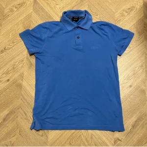 Blå pikétröja från Boss - Säljer en stilren blå pikétröja från Boss. Tröjan har en klassisk krage med två knappar och diskret broderad logga på bröstet. Perfekt för en avslappnad men ändå snygg look.