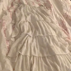 Vit klänning med volanger - Säljer en söt vit klänning med flera lager av volanger och pärldetaljer vid halsen. Klänningen har tunna axelband och en luftig design, perfekt för varma dagar. Den är tillverkad i ett lätt och bekvämt material.