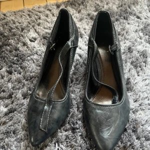 Svarta pumps från Marco Tozzi - Snygga svarta pumps från Marco Tozzi med spetsig tå och klack. Skorna har en dekorativ rem över foten och kontrasterande vita sömmar som ger en stilren look. Perfekta för att lyfta din outfit!