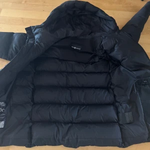 Svart dunjacka från Sail Racing - Säljer en svart dunjacka från Sail Racing. Jackan har en pufferdesign med dragkedja och huva. Perfekt för kalla dagar.