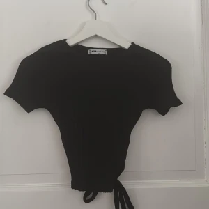 Svart ribbad topp från FB Sister - Snygg svart ribbad topp från FB Sister i storlek XS. Toppen har korta ärmar och en knytning i midjan som ger en cool look. Halv öppen i ryggen, använt typ en gång 🤩🤩