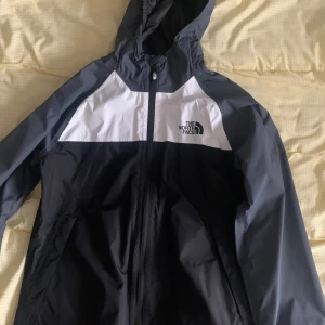 Svart och vit vindjacka från The North Face - JUNIOR STORLEK!! Säljer en stilren svart och vit vindjacka från The North Face. Jackan har en praktisk huva och dragkedja framtill. Perfekt för blåsiga dagar och har en sportig look. Passar bra för utomhusaktiviteter.