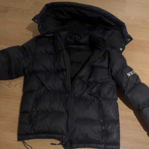 Svart pufferjacka NVLTY - Säljer en svart pufferjacka från NVLTY med huva och dragkedja. Jackan har en stilren design med långa ärmar och är perfekt för kyligare dagar. Den är både snygg och praktisk med fickor framtill. Fräsch jacka knappt använd nästan som ny.