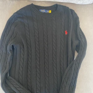 Svart stickad tröja från Polo Ralph Lauren - Säljer en stilren svart stickad tröja från Polo Ralph Lauren med kabelstickat mönster och röd logotyp på bröstet. Tröjan har långa ärmar och ribbstickade muddar. Perfekt för kyliga dagar!