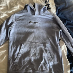 Ljusblå hoodie från Champion - Säljer en snygg ljusblå hoodie från Champion med klassisk logga på bröstet. Tröjan har en stor ficka framtill och en bekväm huva. Perfekt för en avslappnad stil.