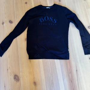 Svart tröja från Hugo Boss - Säljer en stilren svart tröja från Hugo Boss med deras logga tryckt på framsidan. Tröjan har långa ärmar och en klassisk rund halsringning. Perfekt för en avslappnad men ändå snygg look.