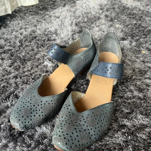 Blå ballerinaskor från Rieker - Snygga blå ballerinaskor från Rieker med perforerat mönster och en dekorativ rem över foten. Skorna har en rundad tå och en liten klack för extra komfort. Perfekta för en stilren look.