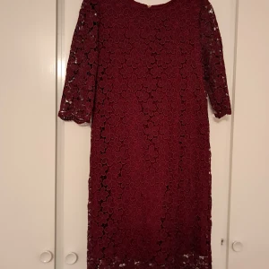 MAYLA (Siden) - Elegant vinröd klänning i spets med blommönster och trekvartsärmar. Klänningen har en dragkedja i ryggen och är fodrad med siden för extra komfort. Perfekt för höst- och vinterevenemang.