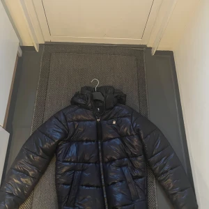 Svart pufferjacka från G-Star RAW - Säljer en svart pufferjacka från G-Star RAW med huva och dragkedja. Jackan har en quiltad design och är perfekt för kyligare väder. Den har praktiska fickor med knappar framtill. Färg mörk blå
