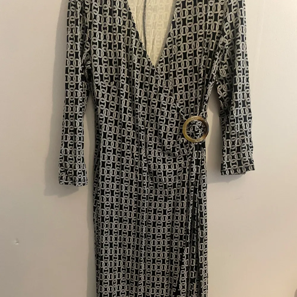 Snygg svartvit omlottklänning från KappAhl med ett geometriskt mönster. Klänningen har en v-ringning och en dekorativ ringdetalj i midjan. Perfekt för en stilren look.. Mekot.