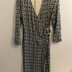 Svartvit omlottklänning från KappAhl - Snygg svartvit omlottklänning från KappAhl med ett geometriskt mönster. Klänningen har en v-ringning och en dekorativ ringdetalj i midjan. Perfekt för en stilren look.