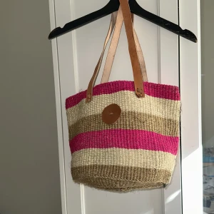 Randig shoppingväska i naturmaterial - Säljer en snygg randig shoppingväska i naturmaterial med rosa, beige och bruna ränder. Väskan har läderhandtag och en praktisk dragkedja upptill. Perfekt för en dag på stan eller stranden!