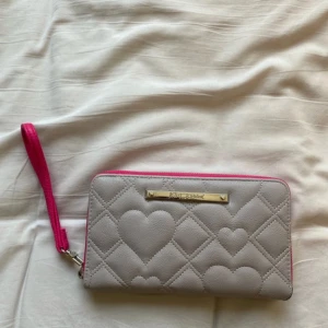 Betsey Johnson Plånbok/Portmonä - Snygg Betsey Johnson plånbok/portmonä med quiltat hjärtmönster i både grått och svart. Den har en praktisk dragkedja och en rosa handledsrem. Insidan är rymlig med flera kortfack och en genomskinlig ficka för ID-kort.