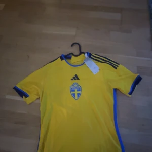 Gul fotbollströja från Adidas - Säljer en gul fotbollströja från Adidas med blå detaljer och Sveriges landslagsemblem på bröstet. Tröjan är i nyskick och har korta ärmar. Perfekt för fotbollsfans som vill stötta sitt lag!