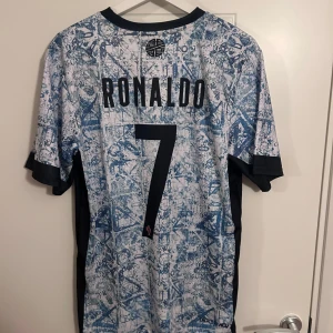 Fotbollströja med Ronaldo 7 - Säljer en unik fotbollströja från Nike med Ronaldo och nummer 7 på ryggen. Tröjan har ett abstrakt mönster i blått och vitt med svarta detaljer. Den är kortärmad och har Portugals landslagsemblem på bröstet.