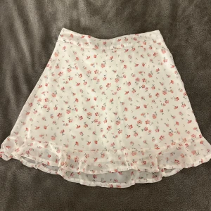 Vit blommig kjol med volang - Söt vit somrig kjol med rosa blommor perfekt till sommaren och dragkedja baktill, använd fåtal gånger och har tyvärr inga bilder med den på. Har en liten defekt som kommer från en säkerhetsnål men inget som syns på. 