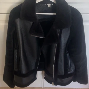 Svart jacka med pälsdetaljer - Snygg svart jacka med pälsdetaljer och dragkedja framtill. Jackan har en klassisk krage och är perfekt för kyligare dagar. Den har en stilren design med en mix av material som ger en lyxig känsla.