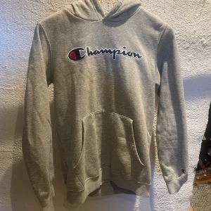 Grå hoodie från Champion - Säljer en grå hoodie från Champion med deras ikoniska logga på bröstet. Tröjan har en klassisk känguruficka och långa ärmar. Perfekt för en avslappnad stil. Den enda effekten är att de är nopprig🙌😀