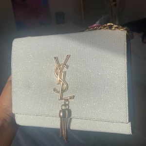 Vit glittrig axelväska med guldkedja - Elegant vit axelväska med en glittrig yta och en stilren guldkedja. Väskan har en dekorativ tofsdetalj och ett ikoniskt logotypemblem i guld på framsidan. Perfekt för att ge en touch av glamour till din outfit.