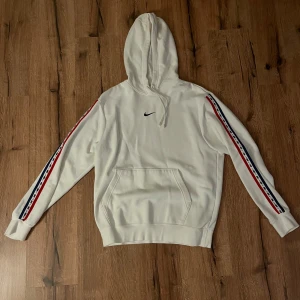 Vit hoodie från Nike - Säljer en stilren vit hoodie från Nike med svarta och röda detaljer längs ärmarna. Den har en klassisk känguruficka och en justerbar huva. Perfekt för en sportig look eller avslappnade dagar. 🏃‍♂️